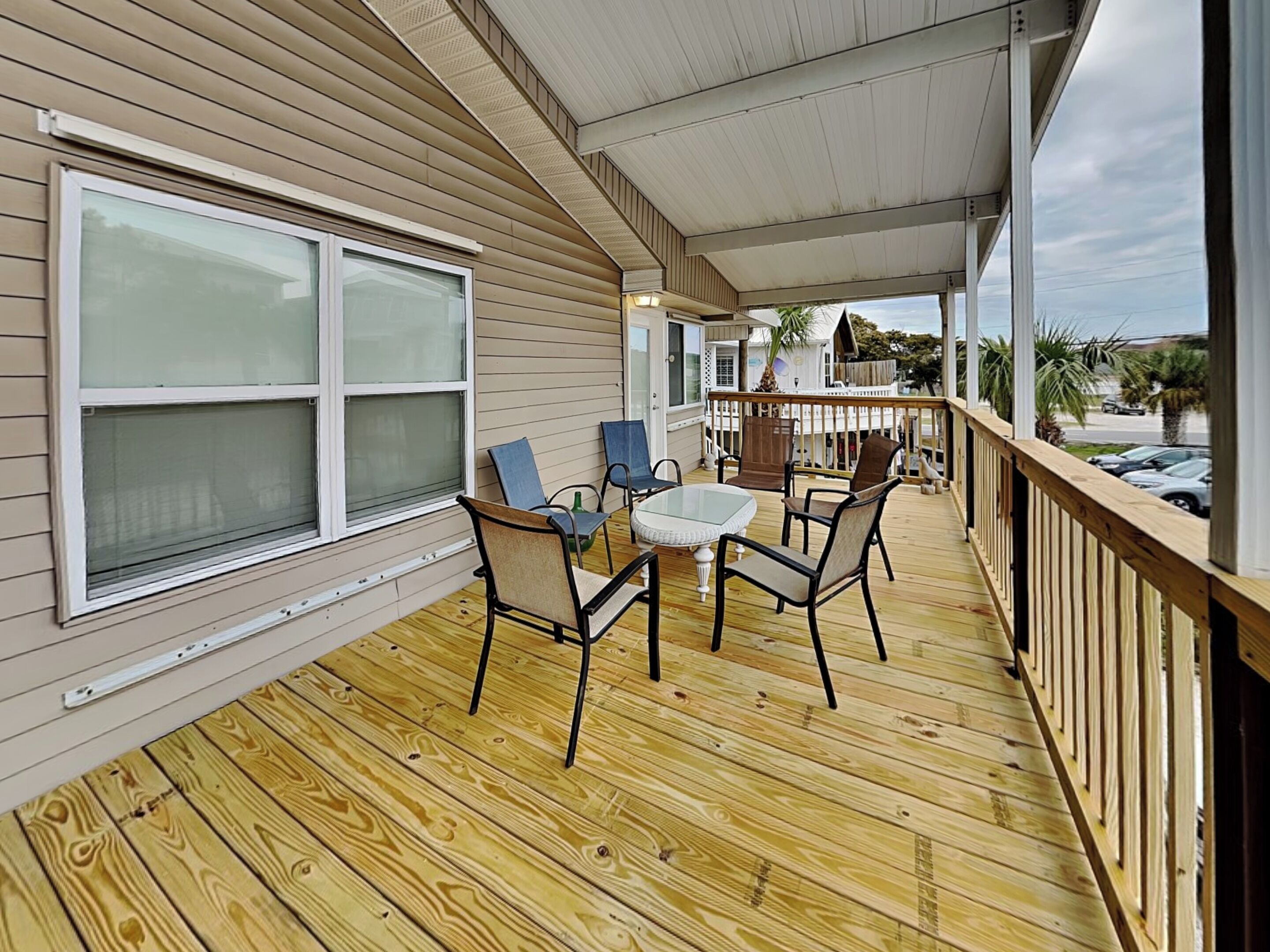 A covered wooden deck with a table and chairs, overlooking a street with parked cars and palm trees.