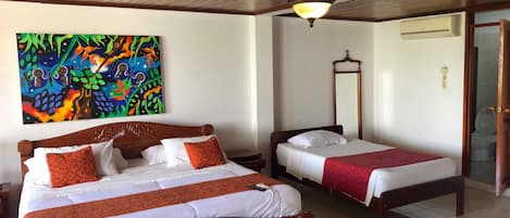 A hotel room with two wooden beds, a colorful abstract painting on the wall, a ceiling light fixture, and a visible bathroom door.