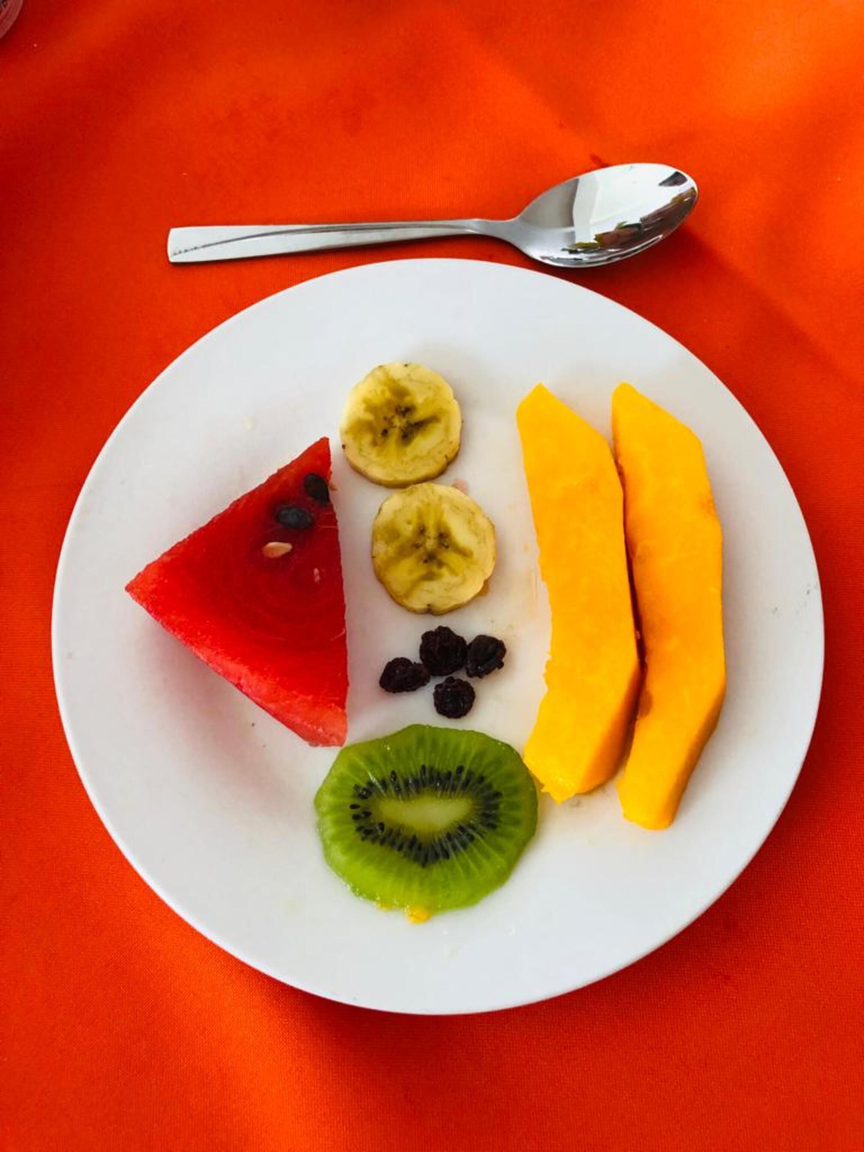 Prato com melancia em fatias, banana, manga, kiwi e frutas secas sobre uma toalha de mesa laranja.