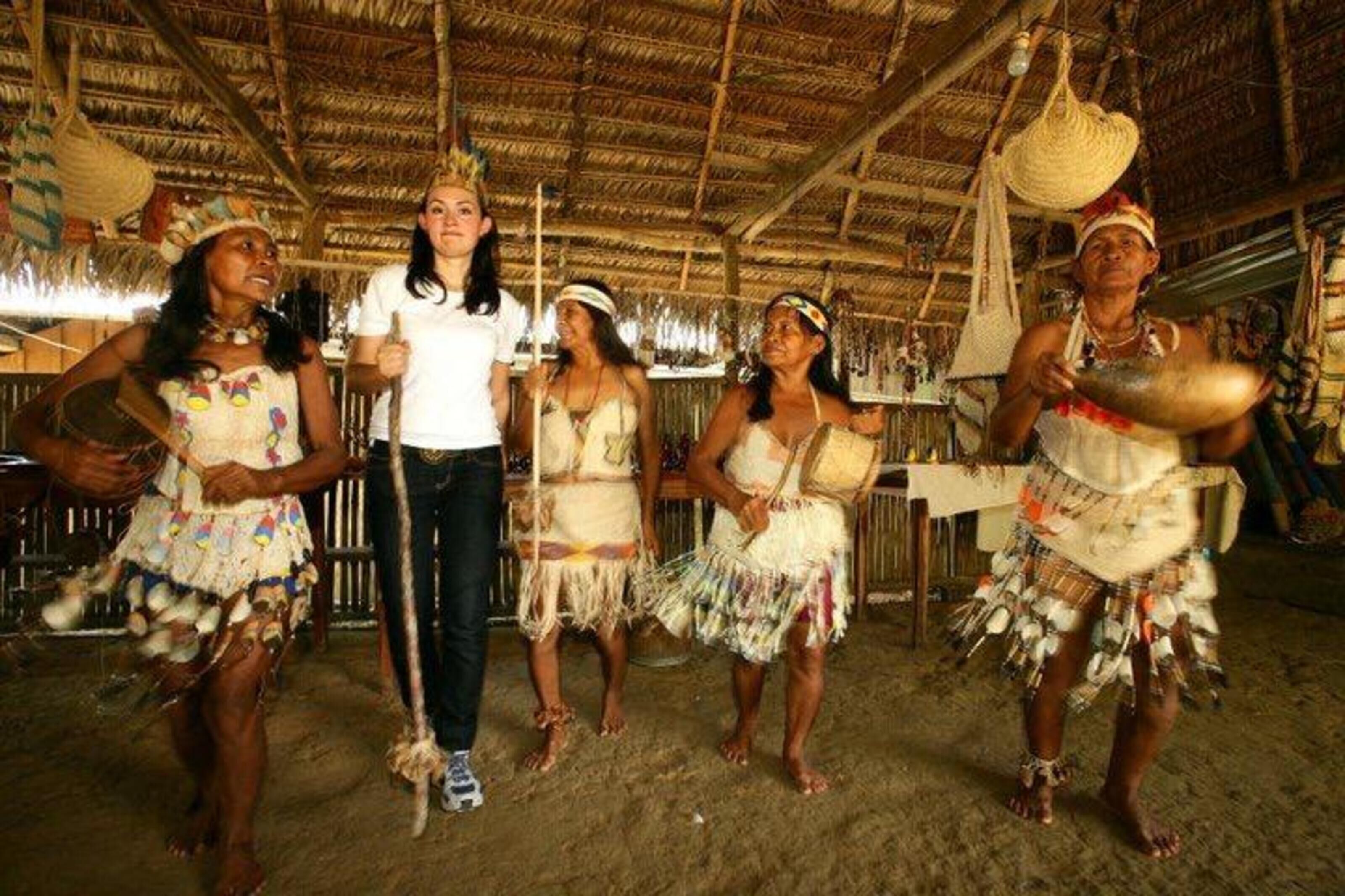 A group of people in traditional attire performing in a rustic indoor setting with a thatched roof and straw flooring.