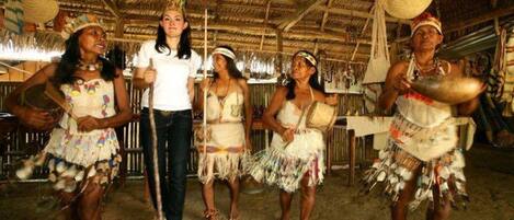 A group of people in traditional attire performing in a rustic indoor setting with a thatched roof and straw flooring.