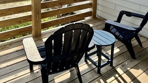 Una terraza de madera con dos sillas negras y una pequeña mesa, con vista a una playa y una casa al fondo.