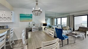 A modern living room with a dining area, a chandelier, and a view of the ocean through large windows. - Emerald Towers by Southern Vacation Rentals (Destin)