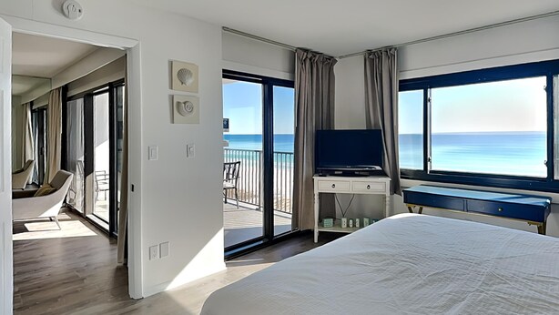 A bedroom with a large bed, a desk, and a TV. There is a balcony with a view of the ocean. - Emerald Towers by Southern Vacation Rentals (Destin)