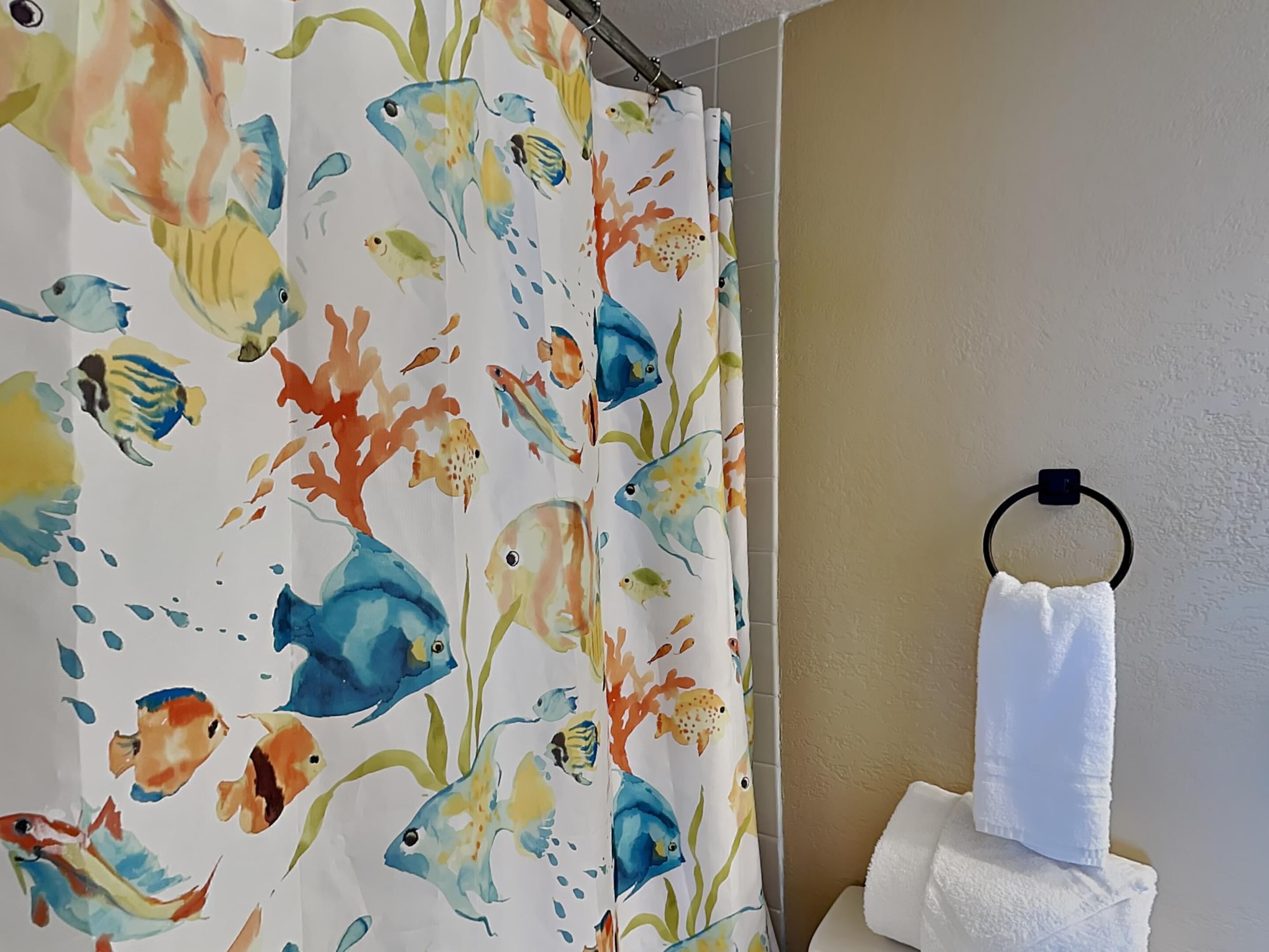 A bathroom with a shower curtain featuring colorful fish and coral patterns, a white towel hanging on a black towel ring, and a beige wall.