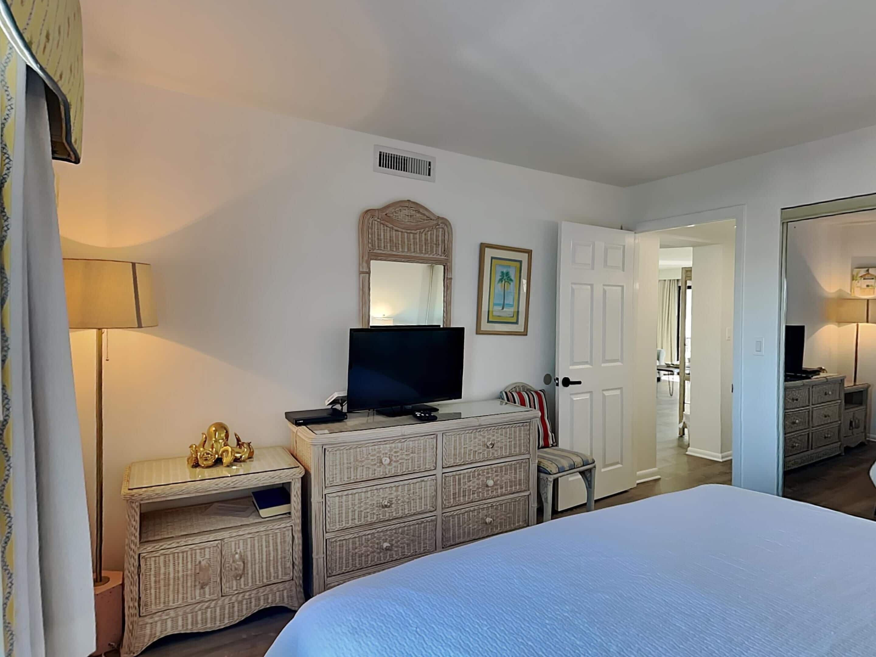 A bedroom with a bed, a television on a dresser, a lamp, and a door leading to another room.