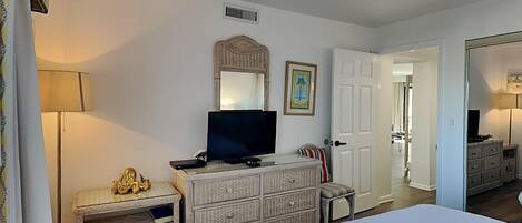 A bedroom with a bed, a television on a dresser, a lamp, and a door leading to another room.