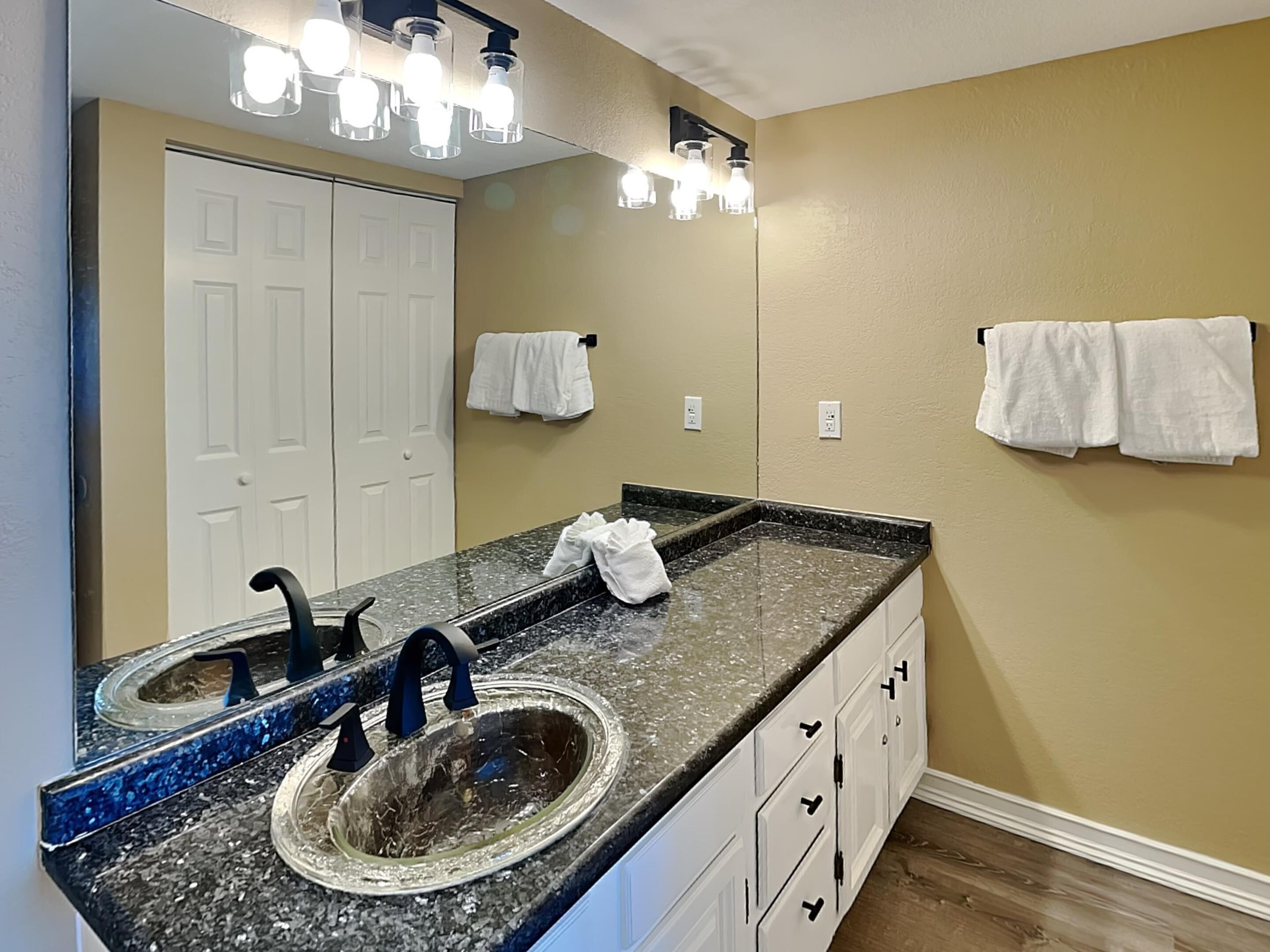 A bathroom with a granite countertop, two sinks, a large mirror, and a towel rack with two towels.