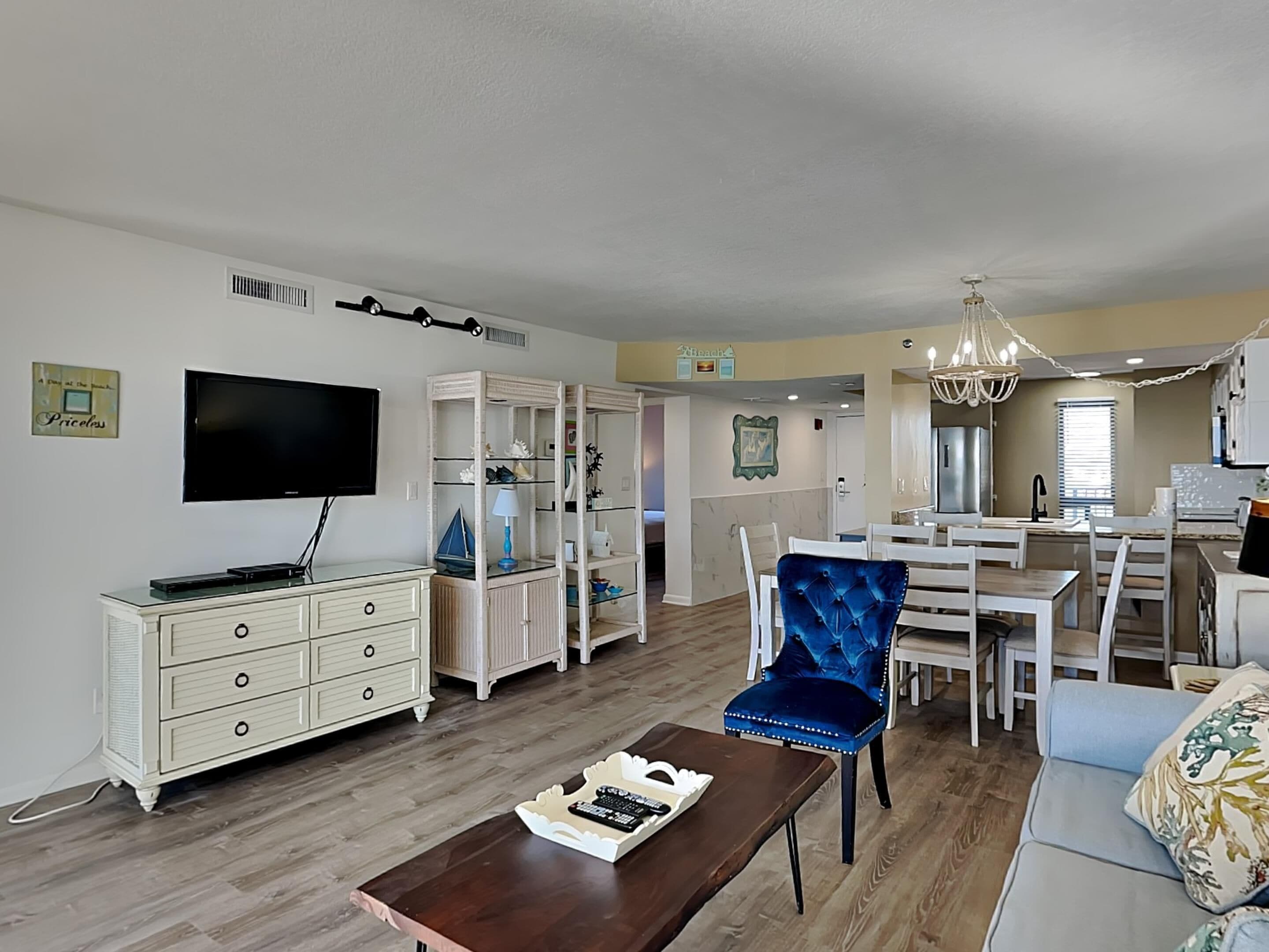 A spacious living room with a television, a dining area with a chandelier, and a kitchen in the background.