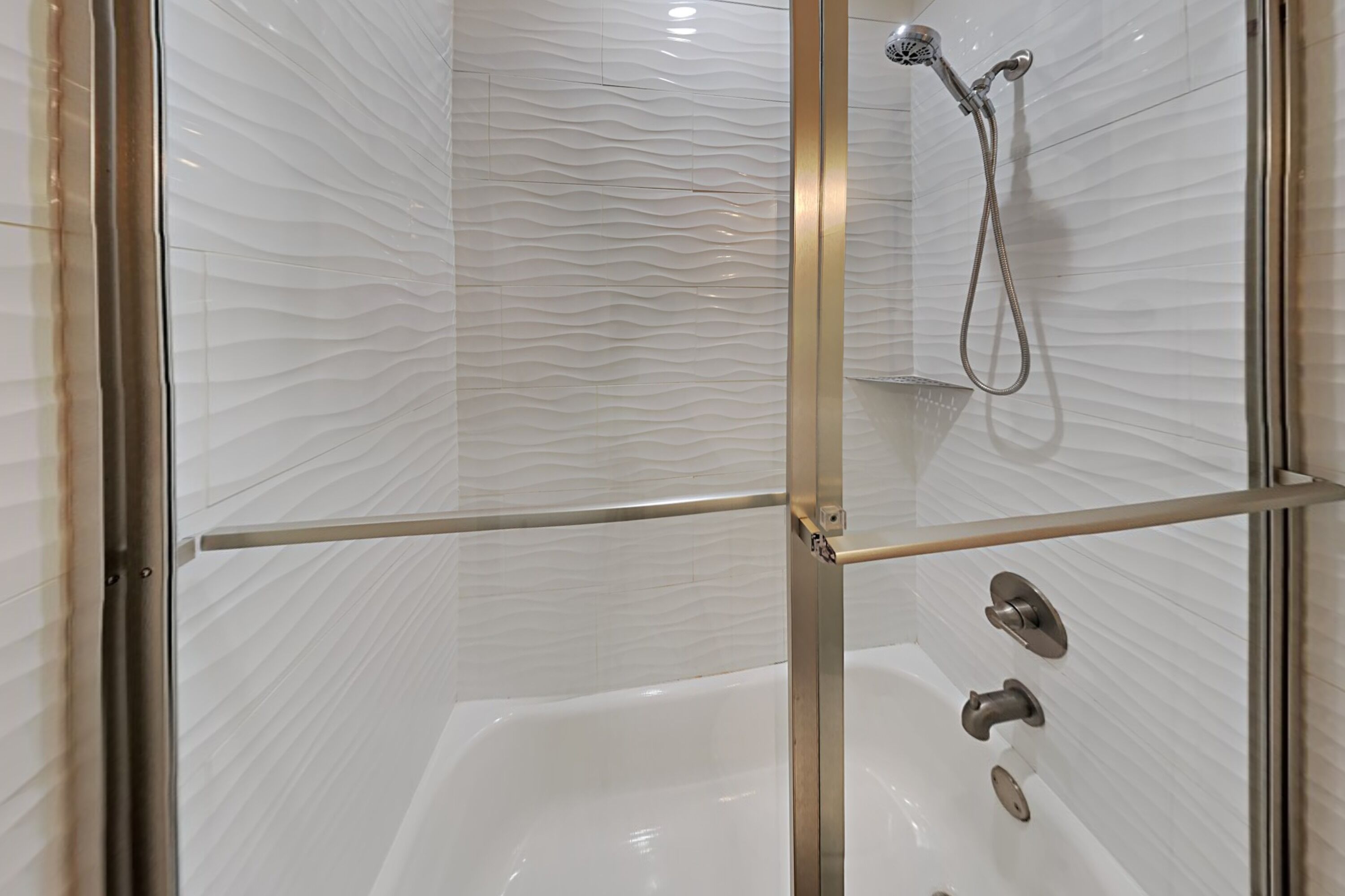 Eine Duschkabine mit Glastüren, einer weiß gestrichenen Wand mit Struktur, einem Handbrausekopf und einem Bedienfeld mit mehreren Wasserhähnen.