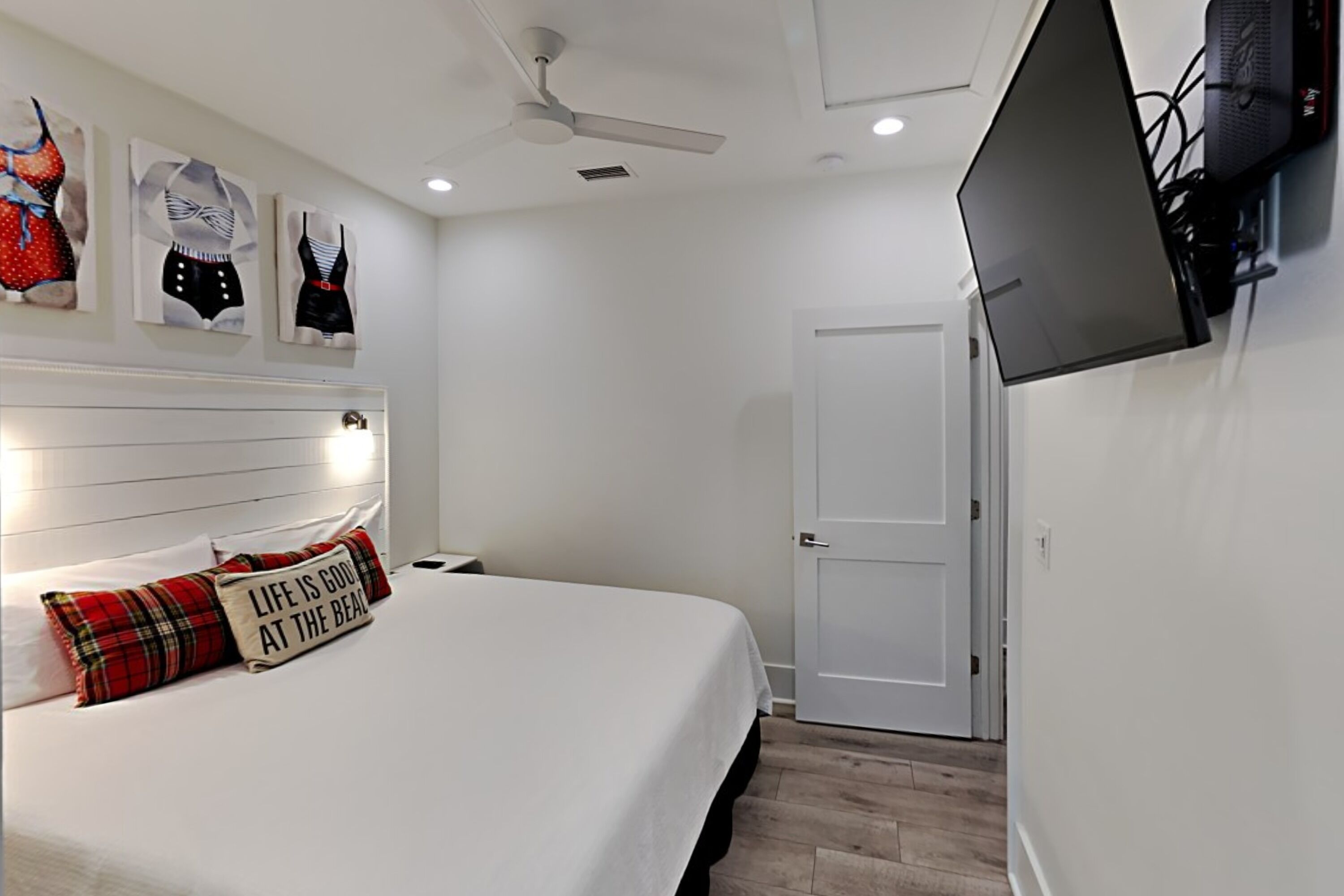 A bedroom with a bed, a ceiling fan, a TV mounted on the wall, and artwork on the wall.