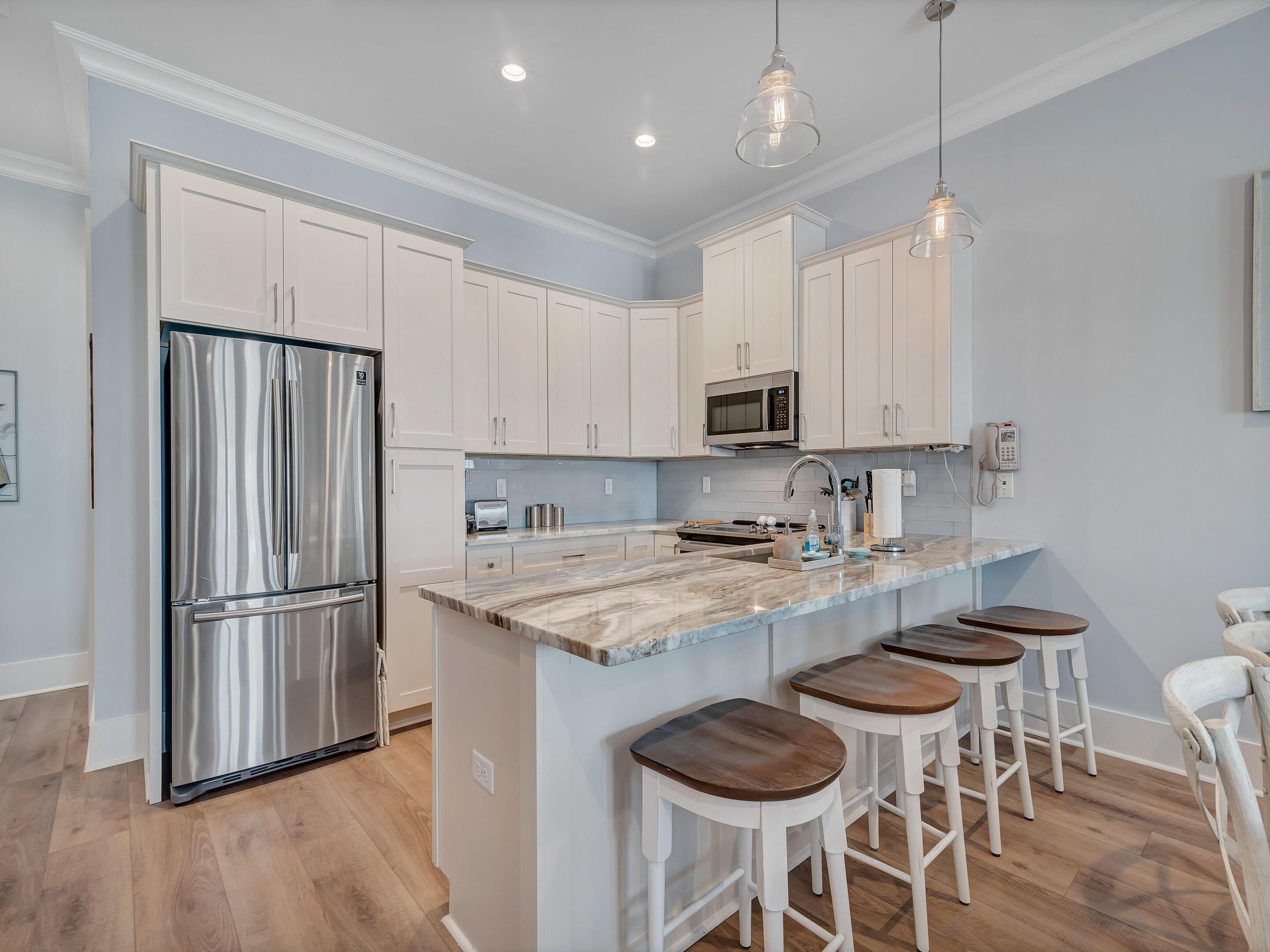 A modern kitchen with white cabinets, a stainless steel refrigerator, a microwave, and a marble countertop. There are four wooden stools around the island.
