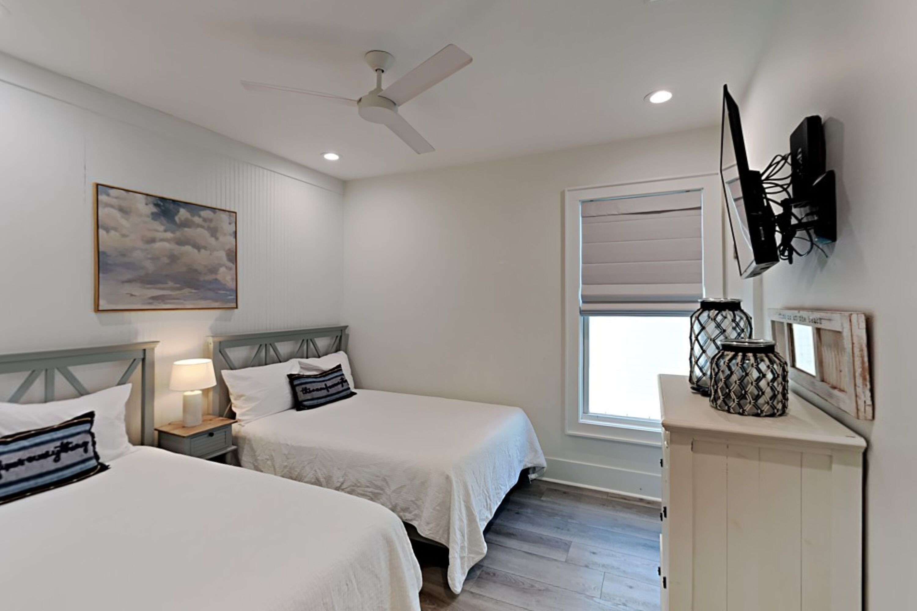 A bedroom with two beds, a ceiling fan, a mounted TV, and a window with blinds.