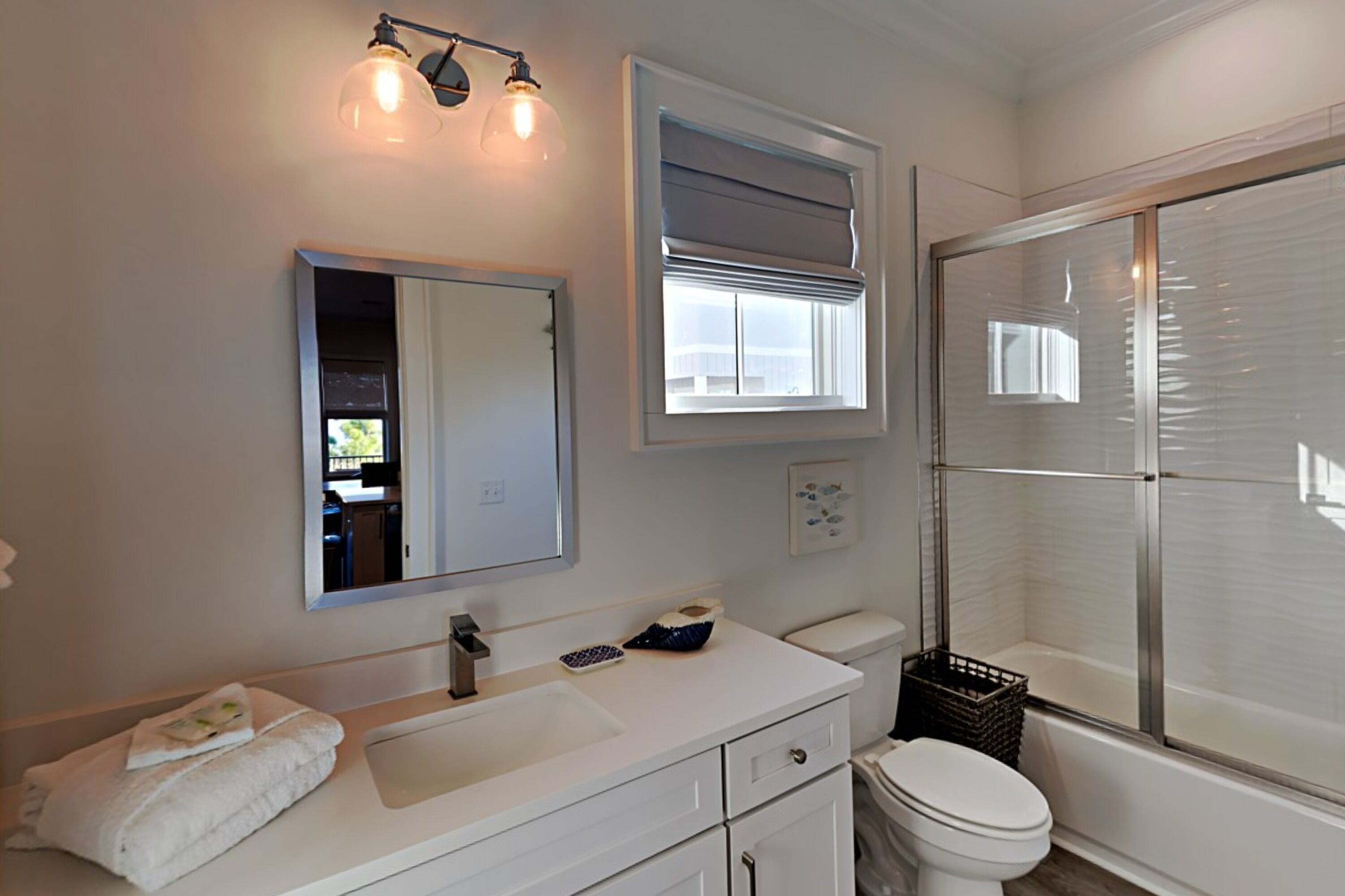 A bathroom with a white sink, mirror, and toilet, a shower with a glass door, and a window with blinds.