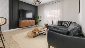 A modern living room with a black leather sofa, a wooden coffee table, a TV mounted on a dark wall, and a large window with sheer curtains. - Stylish 4BR Townhome, Themed Rooms, Private Pool. Near Disney! (Kissimmee)