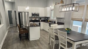 Una cocina moderna con armarios blancos, un refrigerador de acero inoxidable, un microondas y un horno empotrado. Hay una zona de comedor con encimera oscura y sillas blancas.