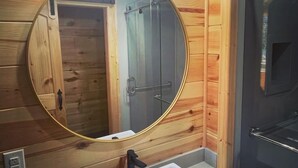 A bathroom with a wooden wall, a round mirror, a white sink, and a shower enclosure. - SUADA-Superior ADA Cabin (Park City)