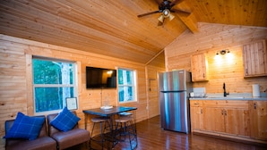 A cabin interior with a wooden interior, a kitchen area, a seating area with a sofa and bar stools, and a television mounted on the wall.