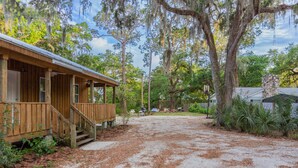 A wooden cabin with a porch, surrounded by trees and a gravel driveway.