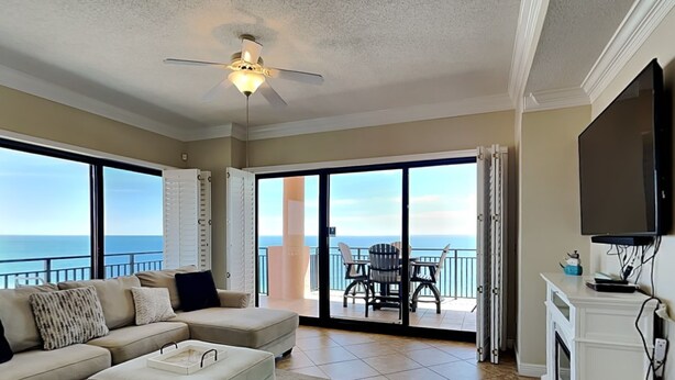 A spacious living room with a large window overlooking the ocean, a ceiling fan, a fireplace, and a flat-screen TV mounted on the wall. - Seachase #1501W by Southern Vacation Rentals (Orange Beach)