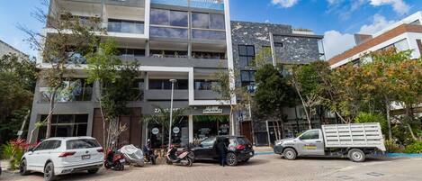 En byggnad med flera våningar och modern design, med en entré på markplan som är markerad med en skylt, och flera parkerade fordon, inklusive bilar och en lastbil.