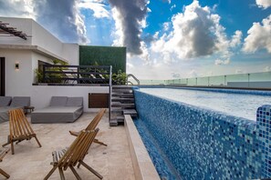 Un área de piscina moderna con un borde revestido de mosaicos, una escalera que conduce a un jardín en la azotea y sillones de madera para relajarse.