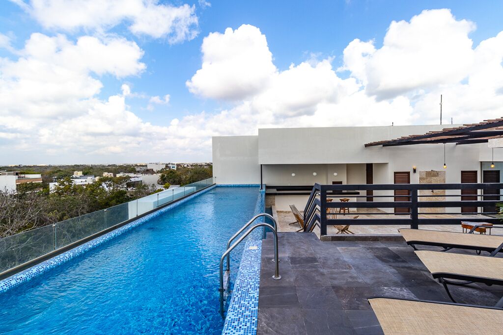 Une piscine sur le toit, avec une eau d’un bleu cristallin, une barrière en verre et une terrasse dotée de chaises longues et de tables.