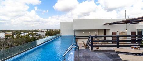 Une piscine sur le toit, avec une eau d’un bleu cristallin, une barrière en verre et une terrasse dotée de chaises longues et de tables.