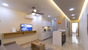 A modern living room with a flat-screen TV, a ceiling fan, and a kitchen area with a bar counter. - Gaia 1-BR Suite (Tulum)