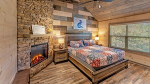 Une chambre confortable avec une cheminée en pierre, un lit recouvert d’une courtepointe, des murs en bois et un ventilateur de plafond.
