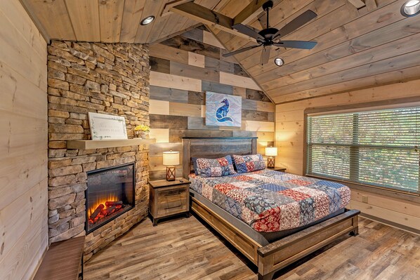 A cozy bedroom with a stone fireplace, a bed with a quilt, wooden walls, and a ceiling fan. - Private Getaway on 3 Acres 5BR Loaded 4King Suites Indoor Pool Theater Game Rm (Sevierville)