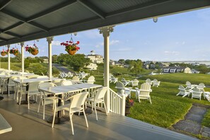 An outdoor patio with white tables and chairs, hanging flower baskets, and a view of a residential area.