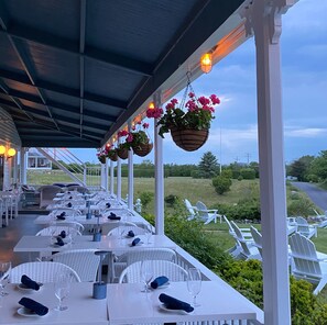 An outdoor dining area with white tables and chairs, hanging flower baskets, and a view of the countryside.