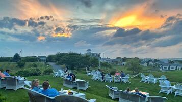 A grassy outdoor seating area with wicker chairs and tables, overlooking a residential neighborhood and a body of water.