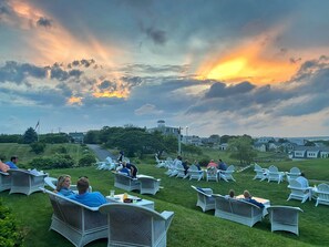 A grassy outdoor seating area with wicker chairs and tables, overlooking a residential neighborhood and a body of water.
