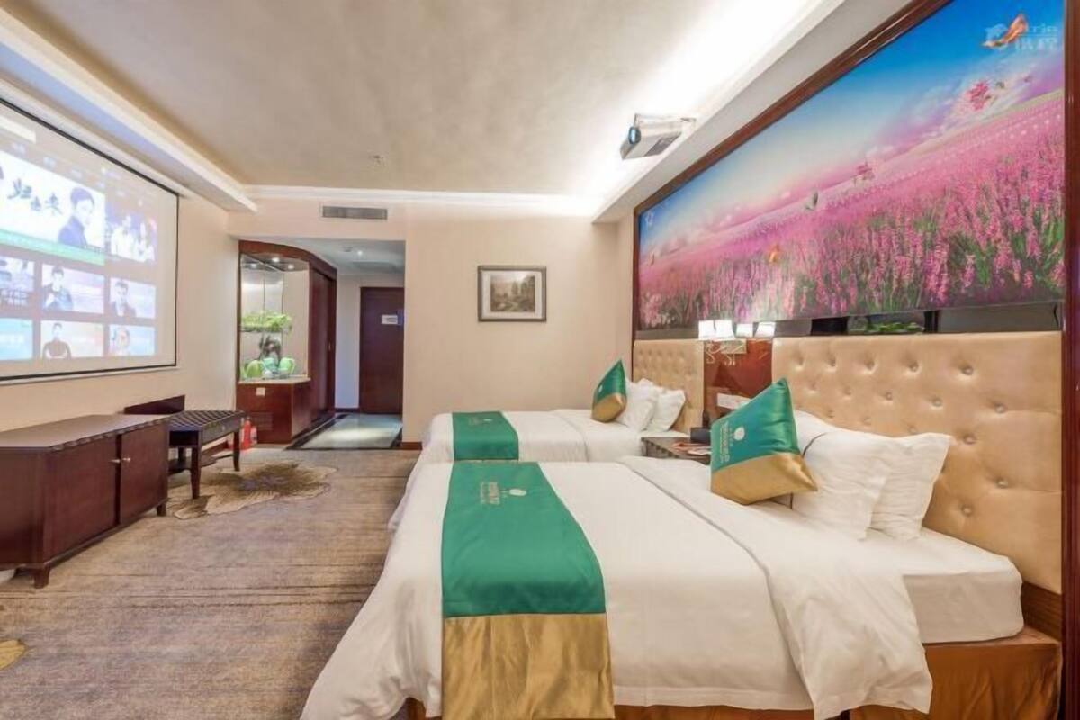 A hotel room with two beds, a large screen, and a painting of a field.