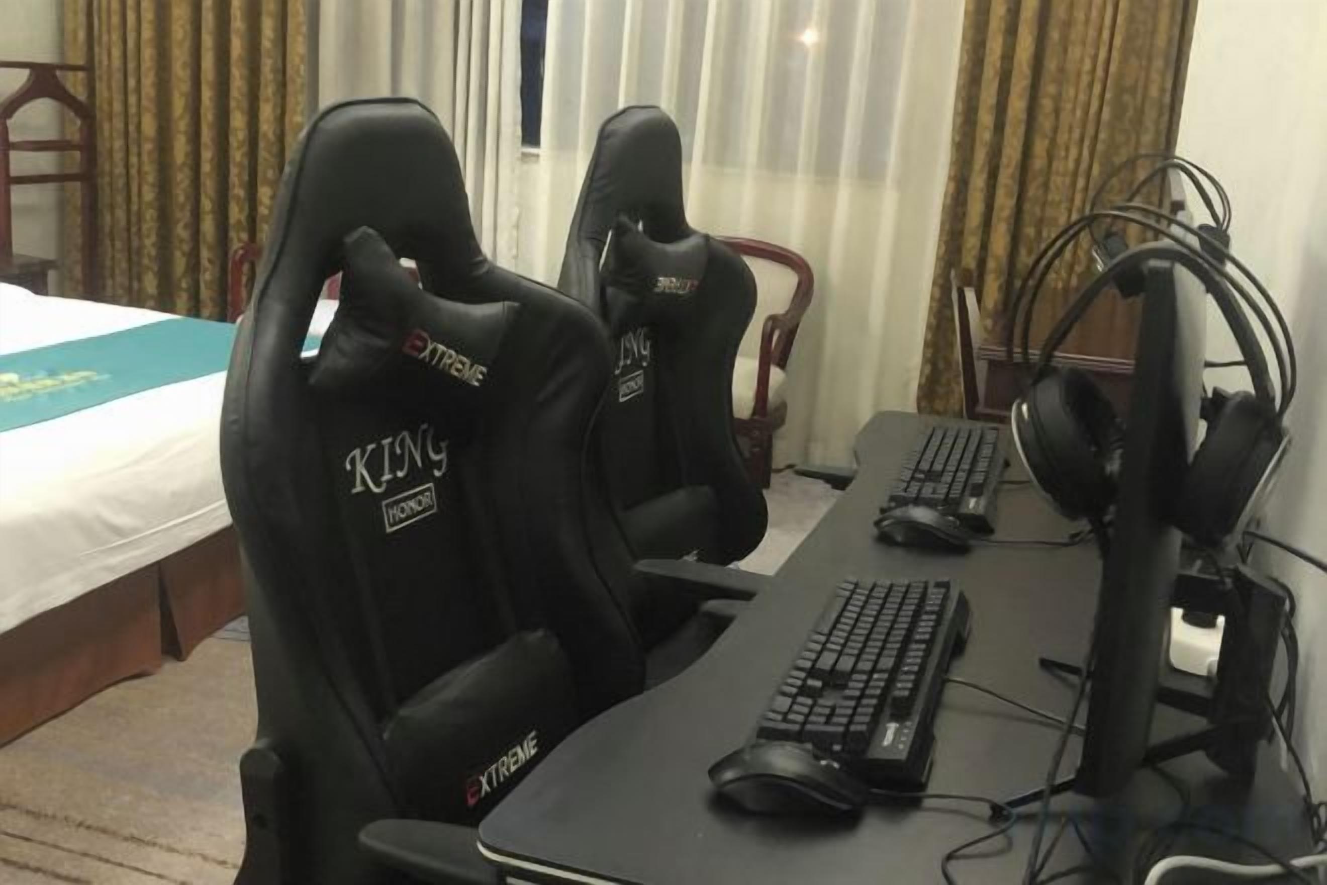 Une chambre d’hôtel équipée de deux fauteuils de jeu portant l’inscription « King », d’un bureau avec un clavier, une souris et un casque, et d’un lit recouvert d’une couverture bleue.