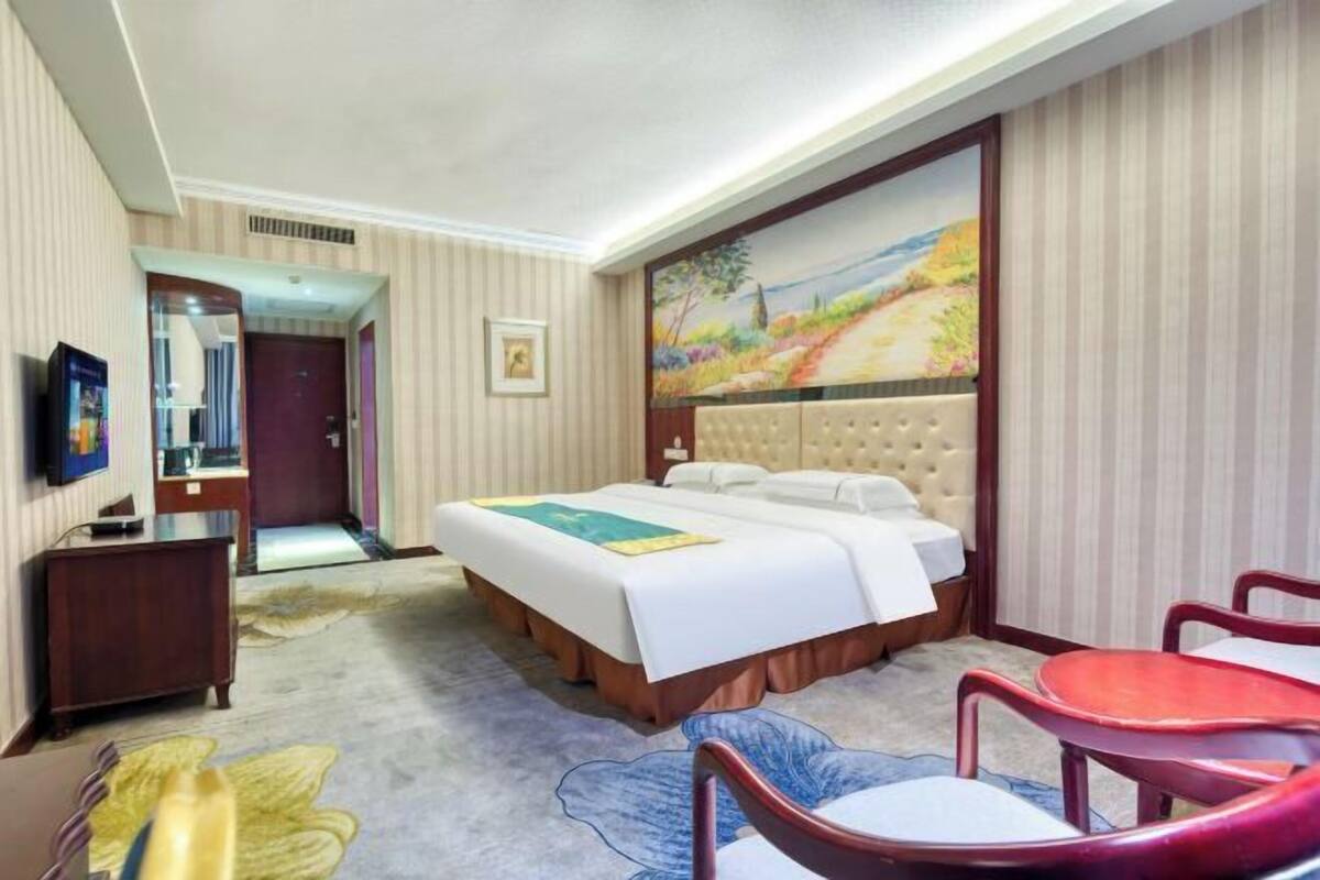 A hotel room with a bed, a desk, a chair, a painting, and a door leading to another room.