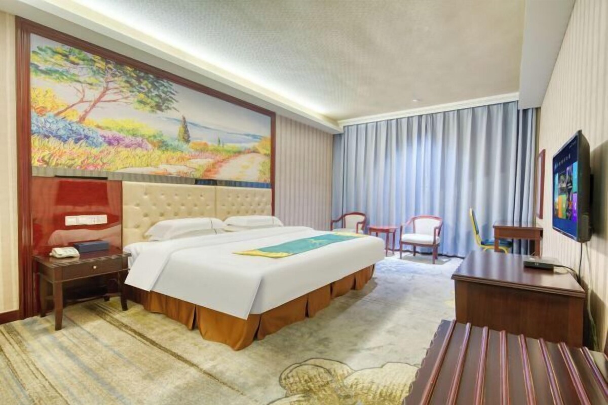 Una habitación de hotel con una cama, un escritorio, una silla, un televisor y un cuadro en la pared.