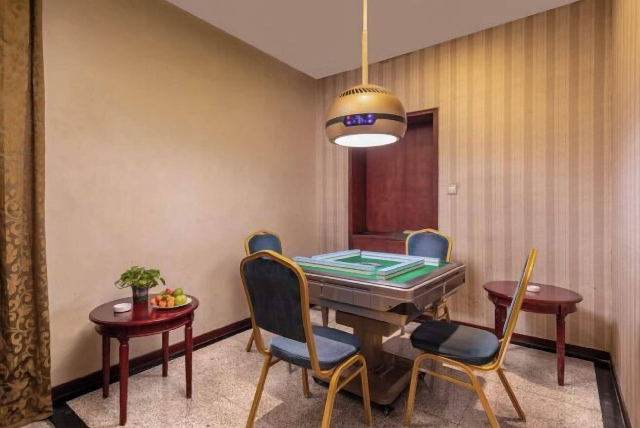 Quarto de hotel com uma mesa de jantar, quatro cadeiras, uma pequena mesa lateral com uma planta e frutas, e um lustre.