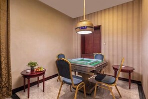 Habitación de hotel con una mesa de comedor, cuatro sillas, una mesita auxiliar con una planta y fruta, y una lámpara colgante.