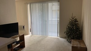 Un salon avec un téléviseur à écran plat posé sur un meuble, un sapin de Noël et une porte-fenêtre coulissante avec des stores.