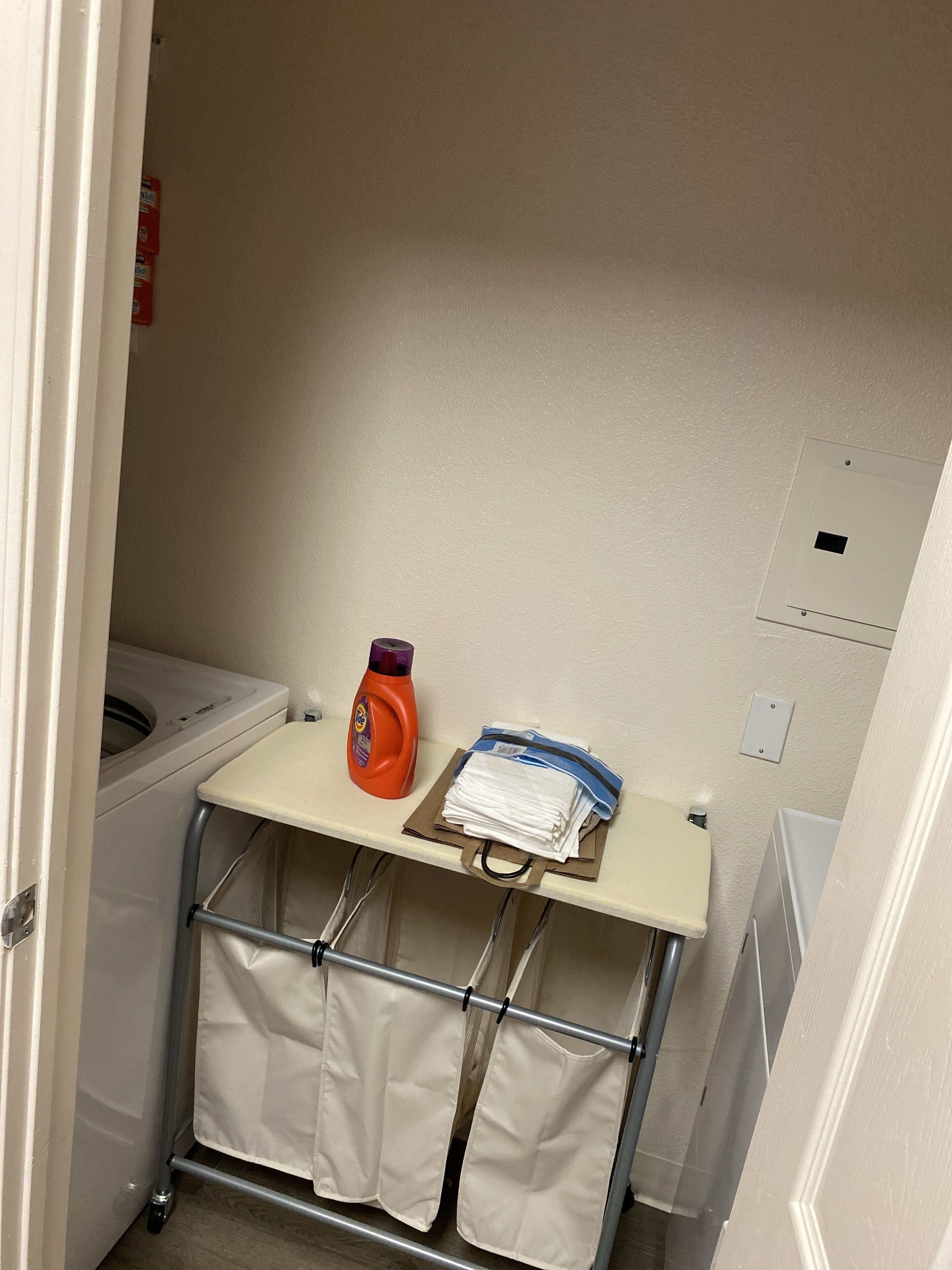 A small laundry area with a washing machine, a detergent bottle, and folded clothes on a shelf.