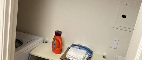 A small laundry area with a washing machine, a detergent bottle, and folded clothes on a shelf.