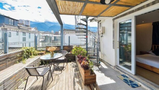 Een balkon met een houten vloer, een tafel met stoelen, potplanten en uitzicht op gebouwen en bergen.