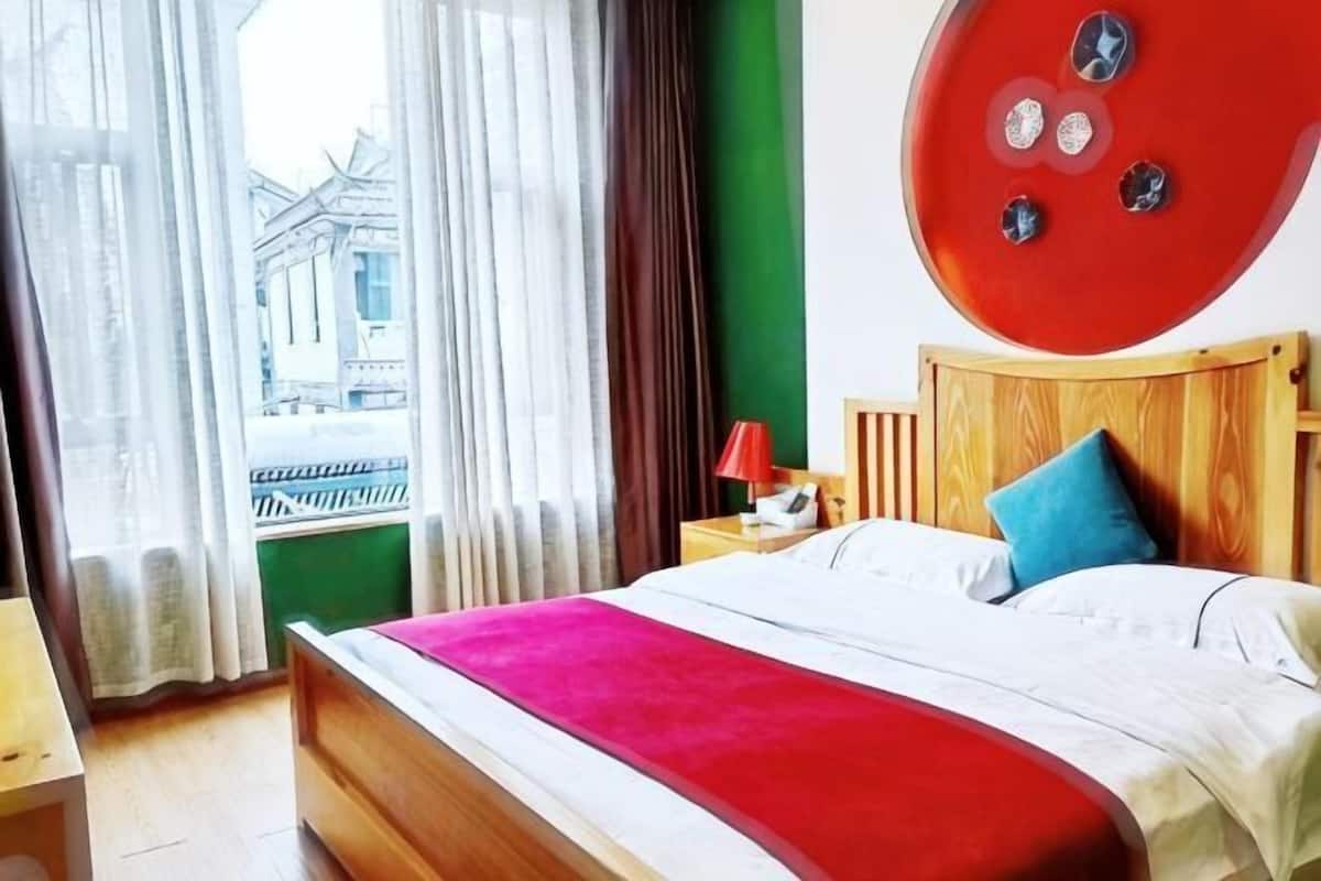 A hotel room with a bed, a nightstand, a red wall decoration, and a view of a building outside.
