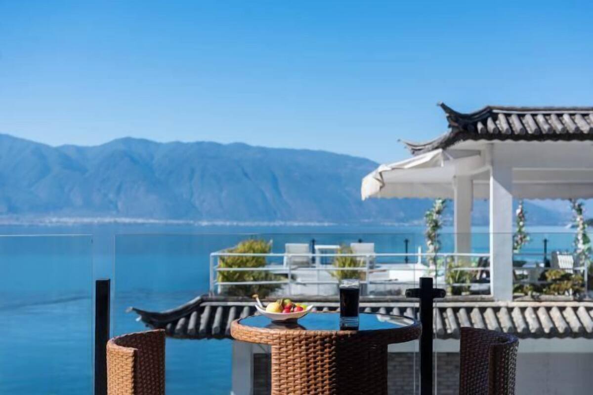 Eine Terrasse mit Blick auf Meer und Berge, mit einer traditionellen, überdachten Struktur, einem Tisch mit Obstschale und einem Glasgeländer.