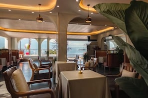 A restaurant with tables and chairs, a view of the sea, and a large plant.