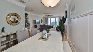 A dining area with a long table, chairs, and a chandelier. There is a television in the background, and a plant is visible on the right side. - Tortuga Shoals (Tierra Verde)