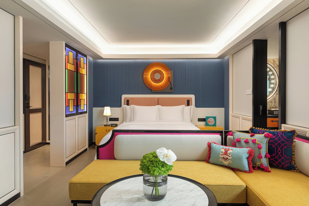 Habitación de hotel con cama, mesita de noche, sofá con cojines coloridos, una pequeña mesa con un florero y una lámpara circular en la pared.